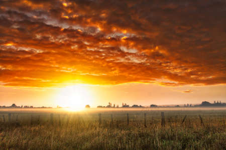 Dramatic vibrant cloudscape  at dawn over mist and fog with a sun flare on the horizon on the farmの写真素材