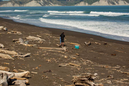 Unknown father and son fishing at a wild rough ocean beach covered in driftwoodの写真素材