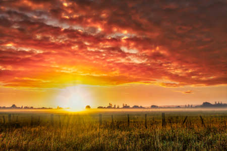 Stunning collared clouds at dawn over the rural agricultural grazing farmland with a little bit of mist and fog and a strong sun flareの写真素材