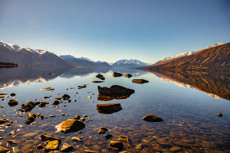 Rocky shore of Lake Ohau with the snow capped Southern Alps reflected in the calm waterの写真素材