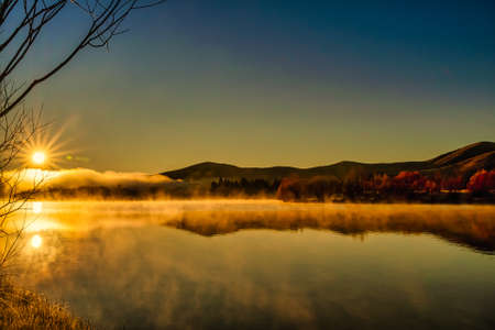 Sun flare and mountain reflections hovering above the water surface at the deserted lake at sunrise in the mist and low cloudの写真素材