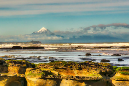 Snow capped volcanic peak of  Mount Taranaki shrouded in cloud viewed from Tongaporutu coastline rock formationsの写真素材