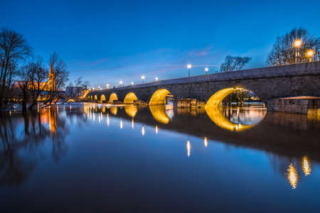 Flood of Danube river in winter 2021 in Regensburg with flooded Jahn Island with view of cathedral and illuminated stone bridge at blue hour, Germanyの写真素材
