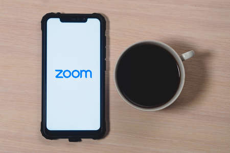 Zoom app logo on the screen smartphone. Zoom Video Communications is a company that provides remote conferencing services. Cup of coffee on a wooden table.のeditorial素材