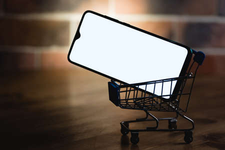 Mockup image of a smartphone with the blank white screen in shopping cart on a wooden table against the background of a brick wall.の写真素材