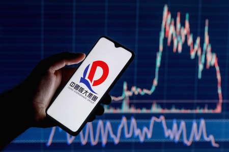 Kazan, Russia - September 22, 2021: The Evergrande Group is the second largest property developer in China by sales. A smartphone with the Evergrande logo in a hand. Stock chart on the background.のeditorial素材
