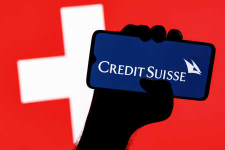 Kazan, Russia - Dec 13, 2021: Smartphone with Credit Suisse bank logo in clenched hand on background of Swiss flagのeditorial素材