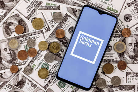 Kazan, Russia - Dec 20, 2021: Smartphone with Goldman Sachs bank logo surrounded by variety of metal coins on background of dollar billsのeditorial素材