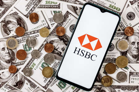 Kazan, Russia - Jan 16, 2022: Smartphone with HSBC bank logo surrounded by variety of metal coins on background of dollar billsのeditorial素材