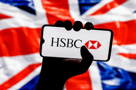Kazan, Russia - Jan 16, 2022: Smartphone with HSBC bank logo in clenched hand on background of British flagのeditorial素材