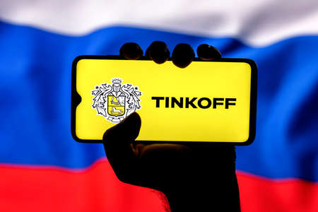 Kazan, Russia - Mar 06, 2022: Smartphone with Tinkoff bank logo in clenched hand on background of Russia flagのeditorial素材