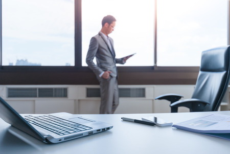 blur business man standing at window using tablet behind working deskの写真素材