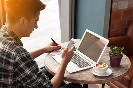 Businessman using smartphone and laptop writing on tablet on wooden table in coffee shop with a cup of coffeeの写真素材
