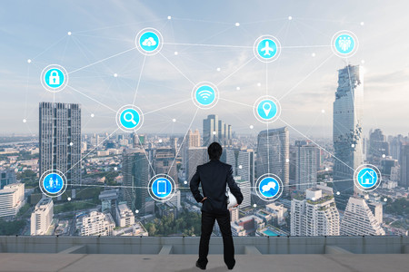 Businessman standing on roof top of skyscrapper looking at icons with cityscape in background,...