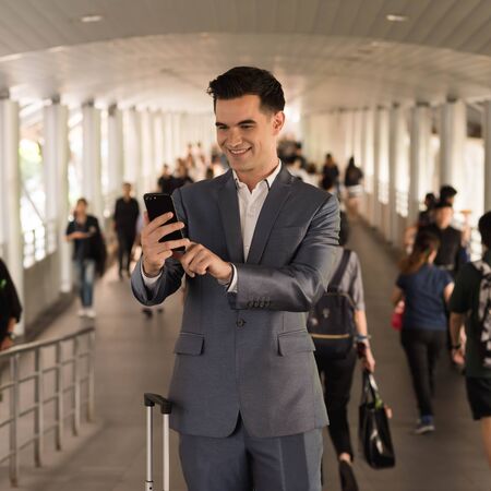 Business man with travel bag use smart phone on the walk way with crowded people in backgroundの写真素材