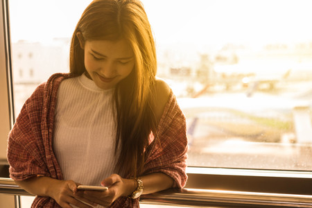Young beautiful woman in airport with travel bag use smartphone with ear phone stand by window wait for flight, world exploration conceptの写真素材
