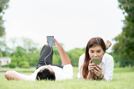 Man and woman on white shirt lie down on grass in public park use smartphone and ear phone together, technology conceptualの写真素材