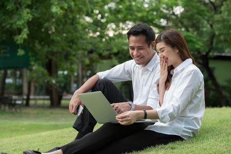 Businessman and woman use laptop in park with happy moment, sit on grass together, laugh at displayed on computer screenの写真素材