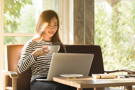 Beautiful young teenage woman freelance work with laptop have coffee at coffee shop in with sun light, freelance lifestyle conceptual, life of coffee loverの写真素材