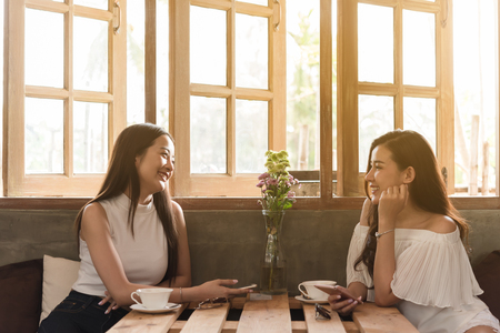 Two teenage women meet in coffee shop use smartphone play social media together in afternoon, life style of new teenagerの写真素材
