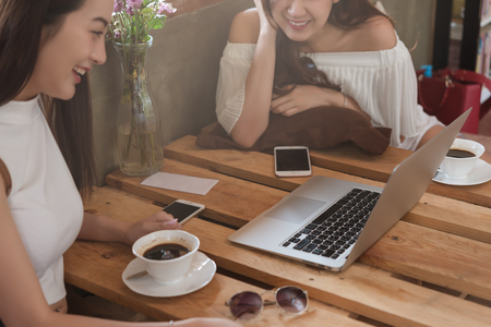 Two teenage women meet in coffee shop use smartphone and laptop play social media together in afternoon, life style of new teenagerの写真素材