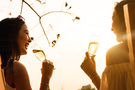 Two beautiful asian women toss white wine of champange in with warm sunset lightの写真素材