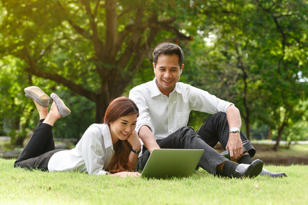 Man and woman in white shirt use smartphone and laptop together in outdoors parkの写真素材