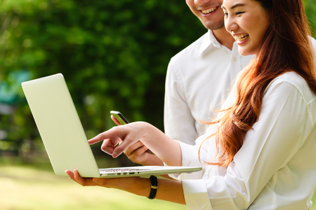 Man and woman in white shirt use laptop and smartphone together in outdoors parkの写真素材