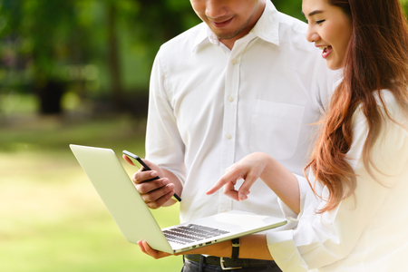 Man and woman in white shirt use laptop and smartphone together in outdoors parkの写真素材