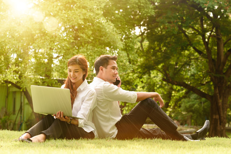 Man and woman in white shirt use smartphone and laptop together in outdoors parkの写真素材