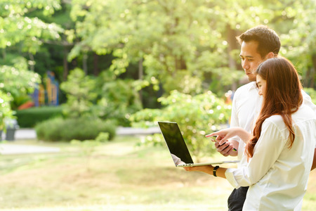Man and woman in white shirt use laptop and smartphone together in outdoors parkの写真素材