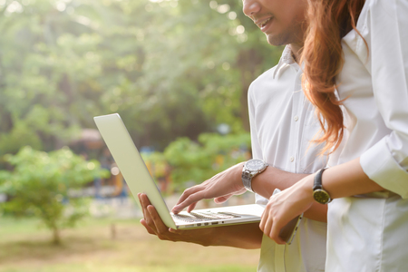 Man and woman in white shirt use laptop and smartphone together in outdoors parkの写真素材