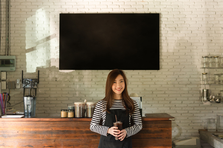 Young woman owner of a cafe stand in front of coffee counter, young entrepreneur conceptual, blank black board on wallの写真素材