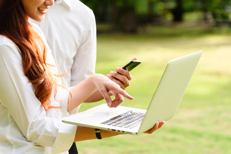 Man and woman in white shirt use laptop and smartphone together in outdoors parkの写真素材