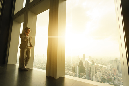 Business man talk via smartphone stand by glass window in tall building with background of cityの写真素材