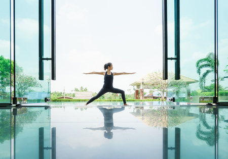 Young asian woman practicing yoga performing Warrior II Pose with reflection on floorの写真素材