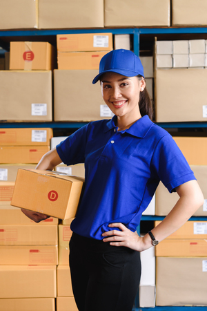 Portrait of woman delivery staff in blue uniform holding parcel box size D in warehouseの写真素材