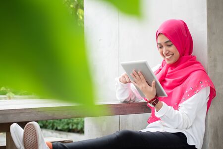 Smiling young muslim woman use tablet play social media wear pink hijab scarf with blur green foregroundの写真素材