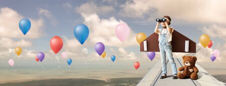 Cute little girl wear diy winged and captain's hat use binocular with her teddy bear buddy imagine that she is flying on the airplane's wingの写真素材