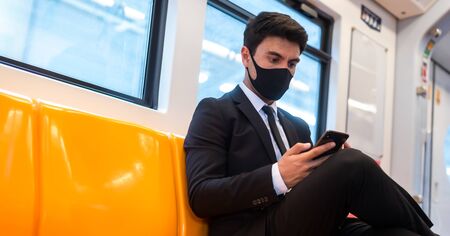 Portrait of confident businessman in black suit wear mask in city finding job during corona crisis using smartphone travelling on empty trainの写真素材