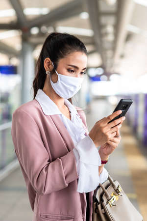 Asian business woman travelling on train in city wearing mask and pink blazer use smartphoneの写真素材