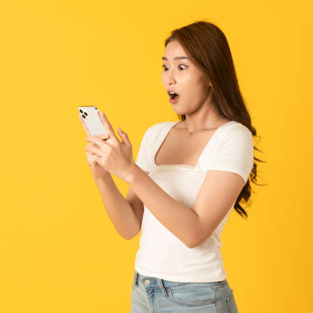 Surprise happy asian beautiful woman isolate on yellow background using smartphoneの写真素材