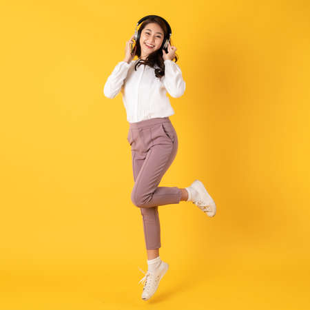 Cute young asian teenage woman using smartphone wearing headphone isolated on yellow background jumpingの写真素材
