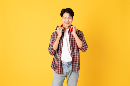 Smile happy asian man wearing scottish shirt with jeans isolated on yellow background using smartphone and headphoneの写真素材