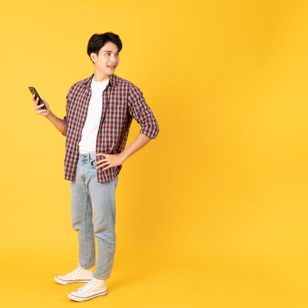 Smile happy asian man wearing scottish shirt with jeans isolated on yellow background using smartphone look to copy spaceの写真素材