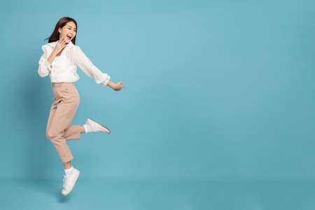 Young beautiful asian woman with smart casual cloth jumping and smiling with excitement looking at copy space isolated on blue backgroundの写真素材