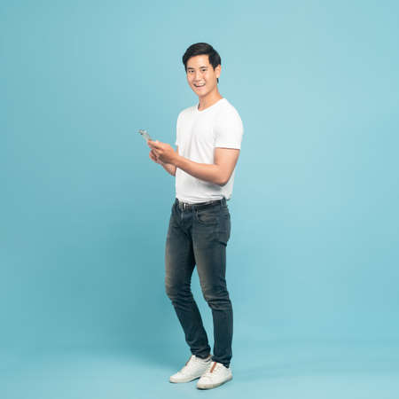 Nice asian guy in white t-shirt and jeans holding smartphone isolated on blue backgroundの写真素材