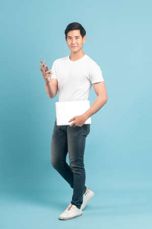Nice asian guy in white t-shirt and jeans holding smartphone and laptop isolated on blue backgroundの写真素材