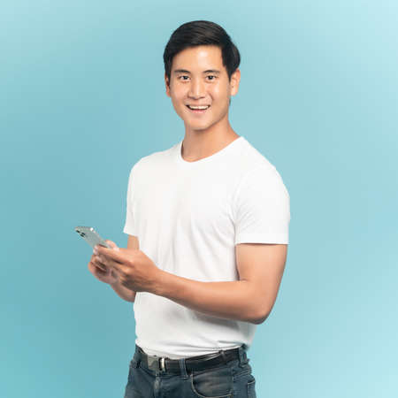 Nice asian guy in white t-shirt and jeans holding smartphone isolated on blue backgroundの写真素材