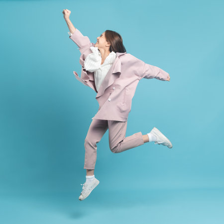 Young beautiful asian woman with smart casual cloth wearing pink coat jumping with excite feeling isolated on blue backgroundの写真素材